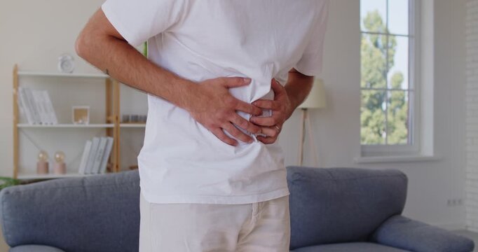 Sick man with abdominal pain holding belly, suffering from stomachache, cramp in stomach or bowel, indigestion or nausea. Person with gastrointestinal problems standing in living room at home