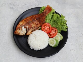 Fried Tilapia or Ikan Nila Goreng. Served on black plate with rice, sambal (chilli sauce), cucumber and tomato slice. Selected focus.