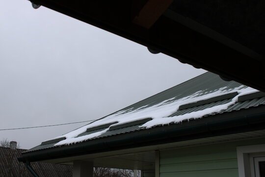 Snow accumulates on a house roof during a cloudy winter day in a quiet neighborhood