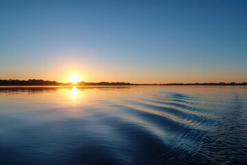 Obraz premium Serene Sunset Over Cityscape Reflected in Deep Blue Water with Golden Light and Clear Blue Sky Illuminating Gentle Ripples on Water Surface