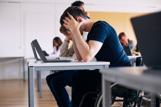 Stressed male high school student sitting on wheelchair with head in hands at classroom