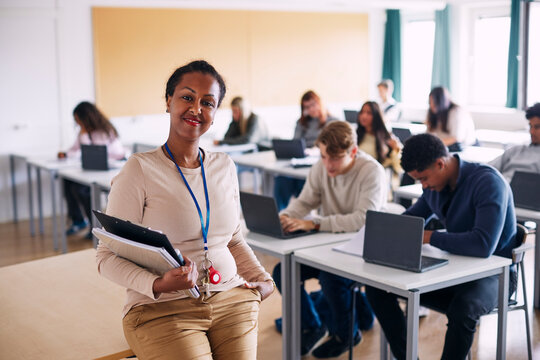 Portrait of confident female educational expert holding books sitting at edge of table in high school classroom
