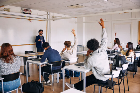Rear view of male students raising hand for asking questions to teacher in classroom at high school