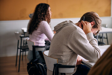 Tensed young male student sitting for educational exam with face down in classroom