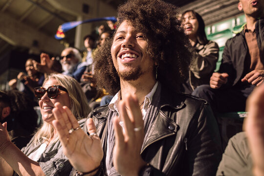 Happy man with curly hair clapping while watching game from stadium stand on sunny day