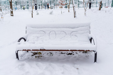 A bench in a winter park is covered with a thick layer of snow, with the number &ldquo;2026&rdquo; playfully written on the surface. The scene evokes festive anticipation, seasonal calm, and holiday atmosphere.