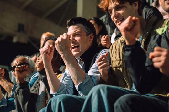Engaged male fan making fist while cheering and sitting in sports stand at sunny day