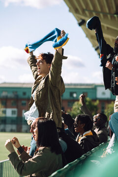 Young man holding sports scarf with raised hands enjoying while cheering from stadium stand at sunny day