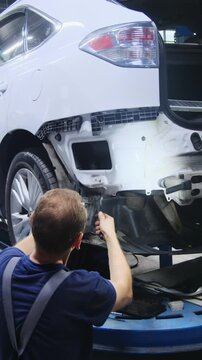 Master works on components of car bumper in large hall. Specialist in overalls modifies surface of modern vehicle standing near wheel