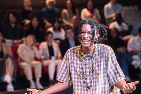 Smiling young male presenter with braided hair gesturing while giving lecture at seminar in theater