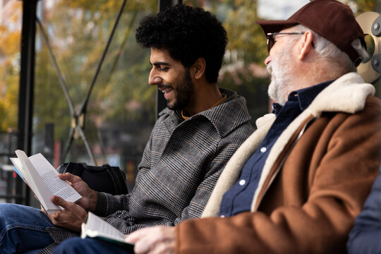 Happy man spending leisure time with father reading book at bus stop on sunny day