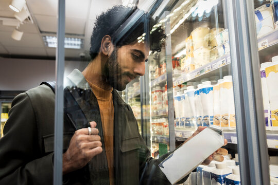 Man reading instructions of milk carton while standing near shelf in supermarket seen from glass door