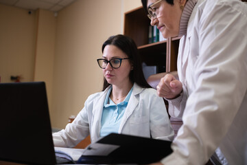 Two professional female doctors in white lab coats collaborating on a laptop in a medical office, focusing on patient records and discussing healthcare information, teamwork and consultation