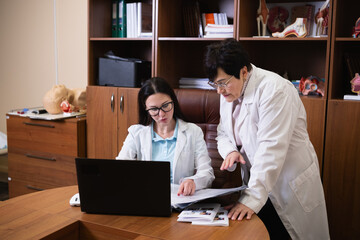 Two female doctors wearing white coats working together, discussing patient data and research findings on a laptop and documents in a healthcare office setting
