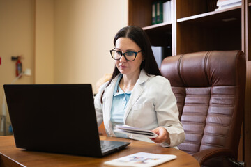 Female doctor wearing a lab coat and glasses sitting at a desk, looking at a laptop while holding documents, representing online medical consultations and digital healthcare