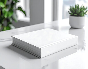 A white book sits on a table next to a potted plant. The book is empty and has a clean, minimalist appearance. The potted plant adds a touch of greenery and life to the scene