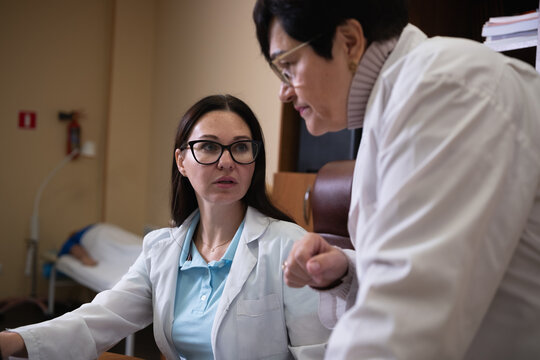 Two female doctors wearing white lab coats and eyeglasses are actively discussing patient information in a medical office, focusing on consultation and healthcare collaboration