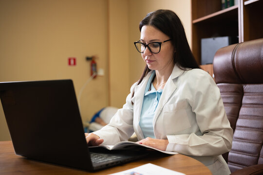 Woman doctor wearing a white lab coat and eyeglasses, sitting at her desk, typing on a modern laptop computer while reviewing data in a clinic consulting room