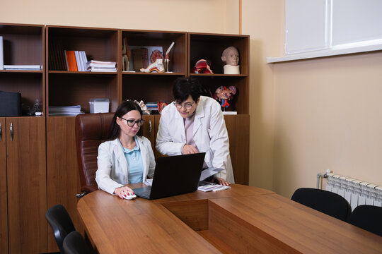 Two female doctors in white coats collaborate in a clinic office, reviewing patient data on a laptop amid anatomical models, focused on diagnosis, research, and care planning - Powered by Adobe