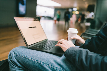 Midsection of female business freelancer holding disposable cup using laptop while sitting at airport terminal