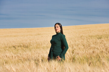 A beautiful, dark-haired teenage Latin girl dressed in green stands among a dry, yellow wheat field on a summer day. In the background, the blue sky.