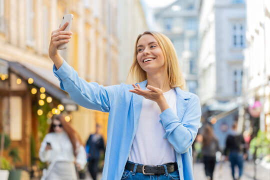 Cheerful woman blogger taking selfie on smartphone, communicating video call online with subscribers or family friends, recording stories for social media vlog, waving hello. Lady on urban city street