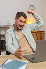 Fototapeta premium Happy young Caucasian man working on laptop netbook shocked by sudden victory, winning, goal achievement, good career news. Excited freelancer guy clenching fists at home office workplace. Vertical.