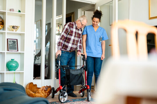 Female healthcare expert assisting senior man walking with rollator at home