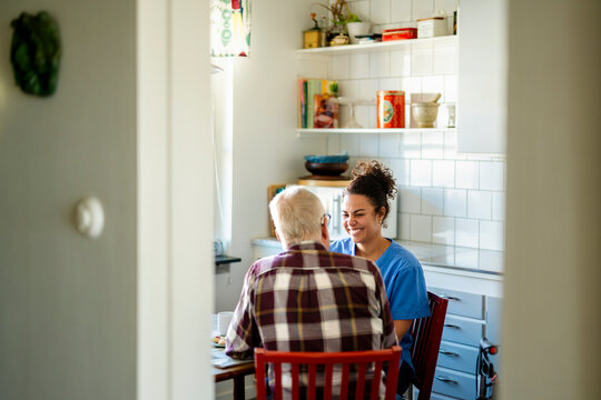 Smiling female home caregiver spending leisure time with senior man sitting in kitchen seen from doorway