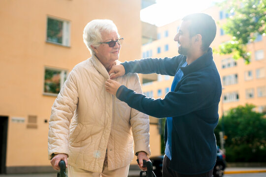 Low angle view of smiling male caregiver buttoning jacket of senior woman at sunny day