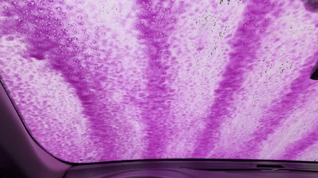 Bright pink detergent spreads across the windshield as an automatic car wash system applies foam,