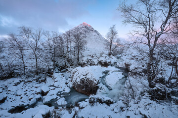 Winter view of Buachaille Etive Mor mountain in Glencoe, Scottish Highlands. Snow-covered landscape with frozen river and soft pink sunrise light illuminating the peak