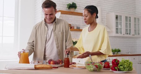Happy diverse couple cooking breakfast together in bright kitchen. Man and woman preparing healthy food with bread, juice, salad and fresh fruits on countertop. Home daily routine and relationships - Powered by Adobe