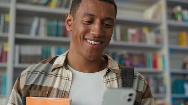 Happy African American man student looking mobile phone smartphone smiling chatting in library technology social media sms online education bookshop university college studying guy male gadget learn - Powered by Adobe