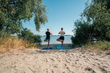 A serene couple practices yoga in tree pose on a sunny beach, overlooking tranquil waters. This outdoor wellness scene embodies balance, mindfulness, and a healthy lifestyle in nature.