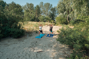 A couple practicing yoga on sandy ground amidst lush green trees and clear blue sky. Healthy lifestyle and outdoor fitness in nature.