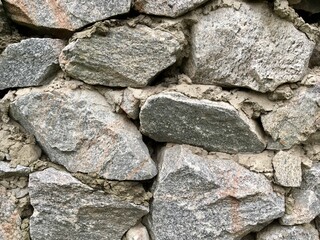 A rustic stone wall, showcasing rough rocks held together by mortar. The stones display a mix of cold light-grey tones, with natural weathering and texture. A horizontal close-up photo.