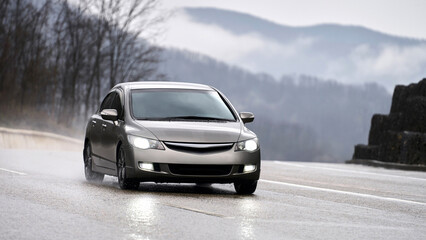 Naklejka premium A silver car with its headlights on a mountain road. It's raining. The headlights reflect off the wet asphalt. Puddles splash from under the tires.