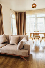Naklejka premium Sofa pillows curtains table floor living home bright interior with modern beige couch and cozy cushions, large windows with sheer drapes and warm hardwood floors in open plan apartment