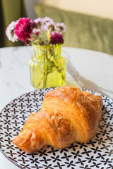 Croissant on floral plate beside vase of fresh flowers in sunlit café interior