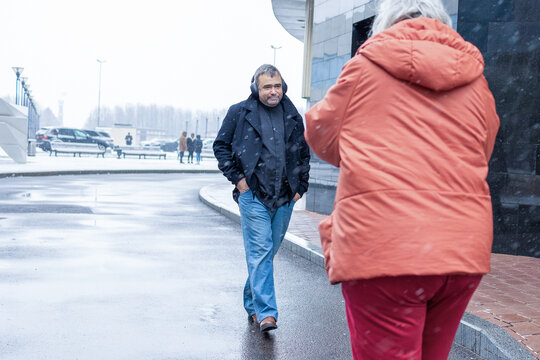 Female photographer in red jacket filming man in headphones listening to music in snowy rainy weather outside modern business center. High quality photo