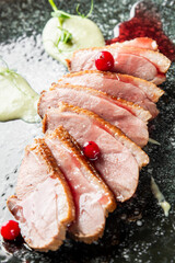 Gourmet sliced duck breast with crispy skin, red berry garnish, and green and red sauces on a dark, elegant plate, close-up