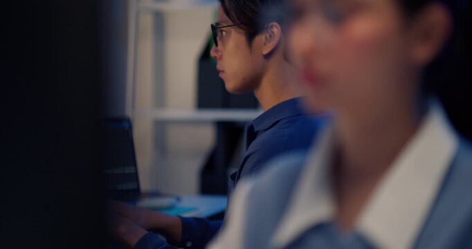 A dedicated young Asian male developer or programmer wearing glasses intently types code on a backlit keyboard. The screen displays lines of programming script in a dimly lit modern office.