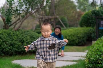 An Asian man and grandmother watching a 16-month-old baby boy run in a park, the grandmother in a blue top and baby in plaid shirt