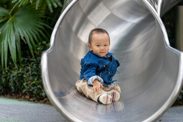 A 16-month-old Asian baby boy in denim shirt playing on a slide outdoors