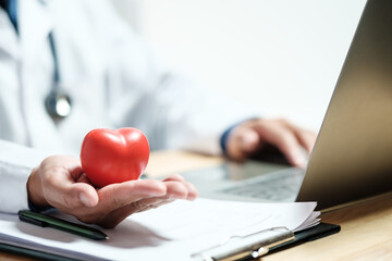 Doctor holding a red heart model while working on a laptop, symbolizing cardiology, healthcare insurance, and online medical consultation. Concept for digital diagnosis and patient record review.