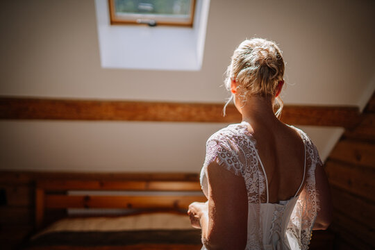 Bride in elegant white dress stands gracefully in sunlit room, showcasing intricate lace details, with wooden beams and soft natural light creating a serene atmosphere - Powered by Adobe