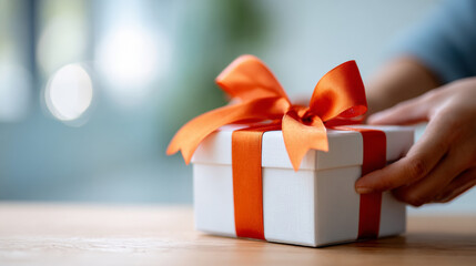 A white gift box with a red ribbon and orange bow is being opened by someone's hands on top of a wooden table