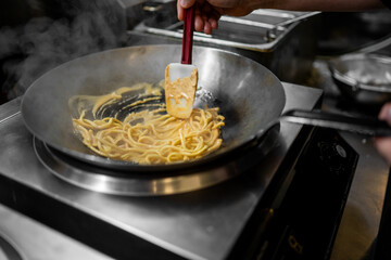 Chef cooking creamy pasta in a large, steaming wok using a red and white spatula in a professional, stainless steel kitchen.