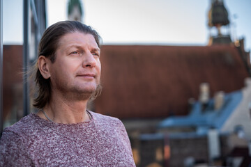 Middle-aged man with long hair stands outdoors, gazing thoughtfully into the distance, with a blurred urban background showcasing rooftops and architectural details